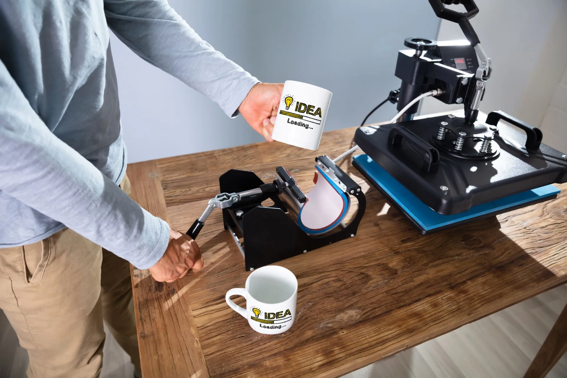 Printers-Proline-Viroqua-025-1889h Person using a mug heat press machine to customize white ceramic mugs with "IDEA Loading..." design, showcasing custom signage and promotional products printing services.