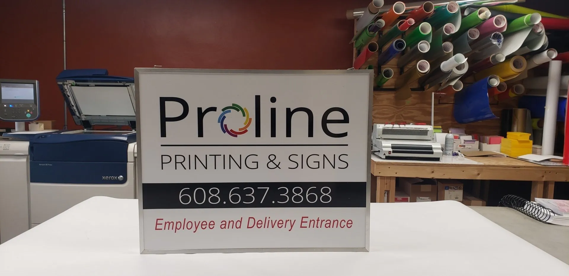 proline+sign+1-1920w Proline Printing & Signs employee and delivery entrance sign with contact number, surrounded by printing equipment and rolls of colorful vinyl in a professional printing and signage workspace in Viroqua, WI.